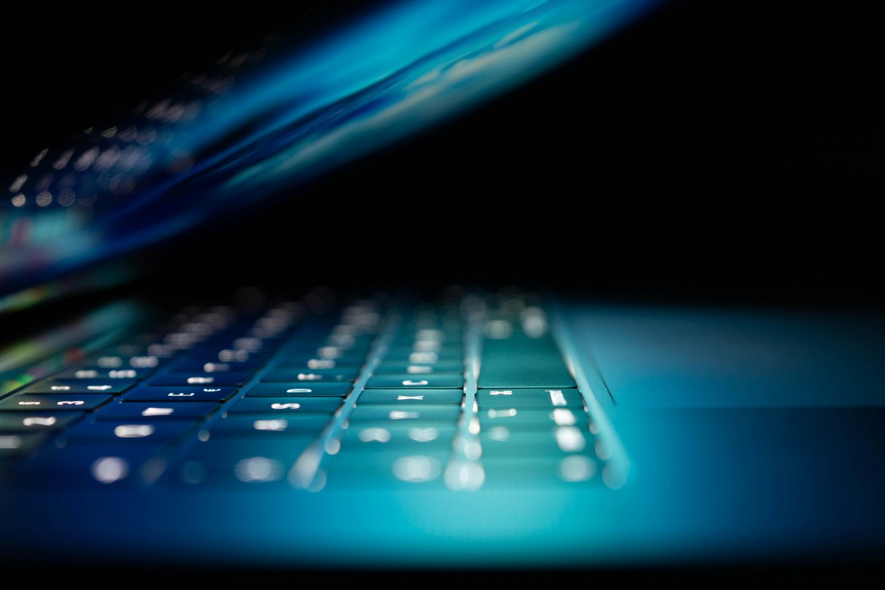 Close-up view of a laptop keyboard with shallow depth of field, showing white backlit keys in the foreground that gradually blur toward the background. The image has a moody blue and teal color scheme with dramatic lighting, creating an atmospheric tech-focused aesthetic.
